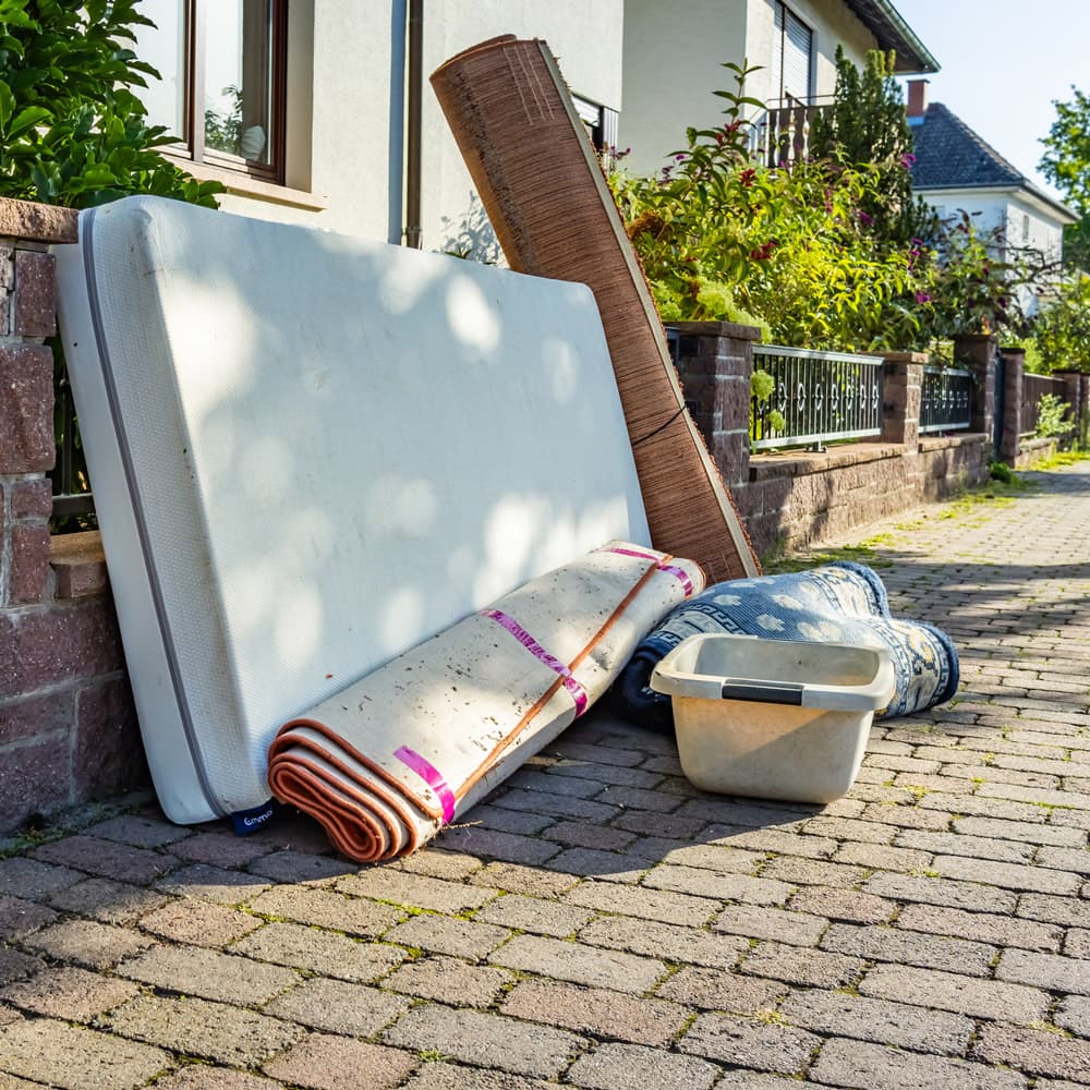 Junk including a mattress, rug, plastic container, and large floor mat sit on the sidewalk in front of a home