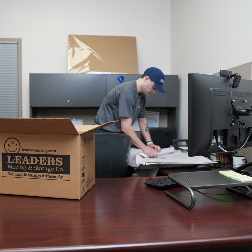 A Leaders Moving & Storage Co. team member carefully packs items in a commercial office with a Leaders box sitting on a computer desk in the foreground for an office relocation