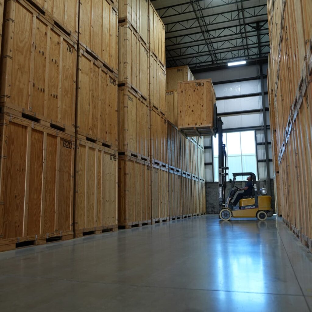 A yellow forklift carefully unloads a large crate from a tall stack of commercial storage crates in a warehouse