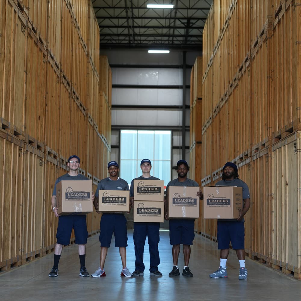 Five Leaders Moving & Storage Co. team members stand proudly holding boxes in between storage crates stacked tall in a warehouse
