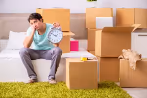 Frustrated renter holding an alarm clock while waiting among moving boxes, illustrating apartment move delays